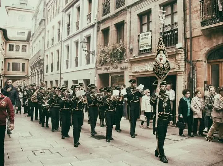 La agrupación desfila tocando en formación por una calle del casco antiguo de Oviedo, con el banderín al frente y los vecinos mirando desde la acera.
