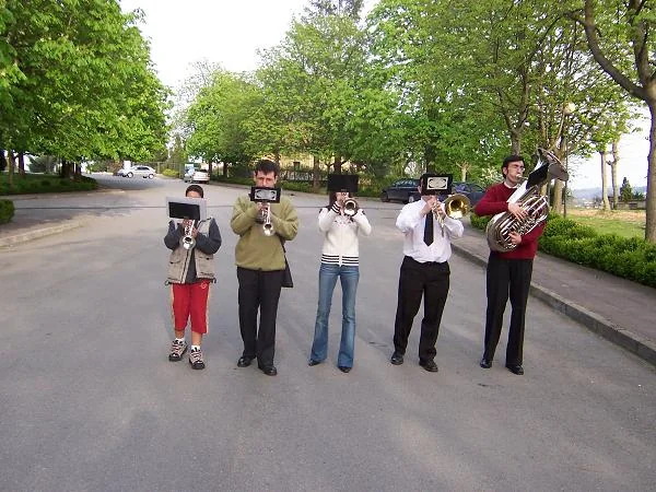 Cinco músicos de paisano, con vaqueros y camisa, tocando con sus atriles en medio de una carretera arbolada.